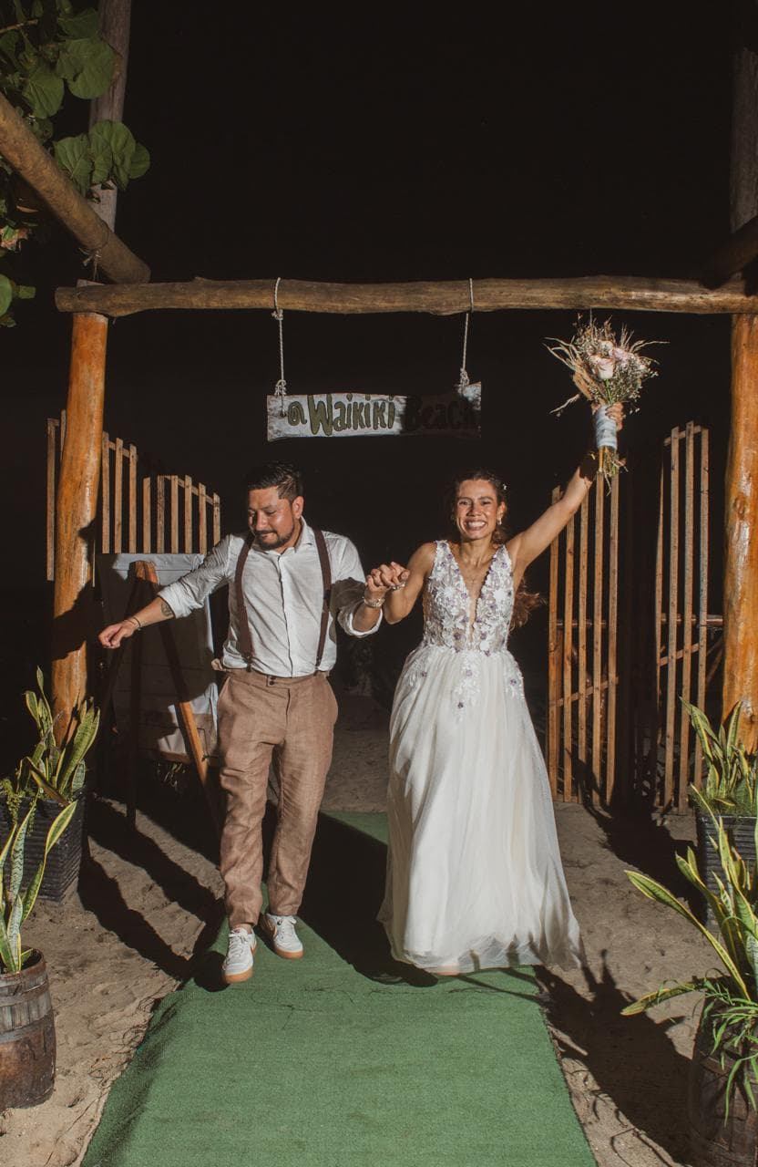 A joyful couple walks hand in hand under a sign that reads "Waikiki Beach," celebrating their wedding at night.