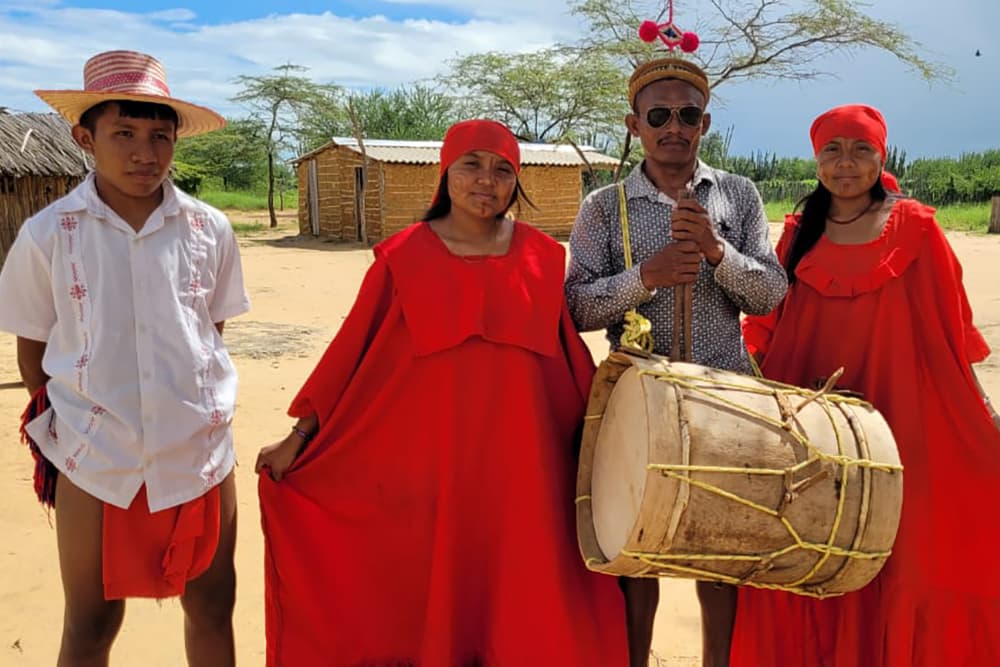 Four individuals dressed in traditional red attire pose together, with one holding a drum, in a rural setting.