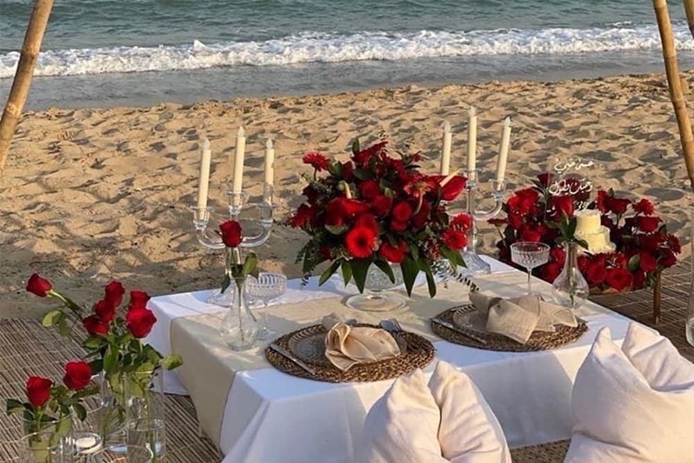 A romantic beach setup featuring a elegantly decorated table with red roses, candles, and a small cake.