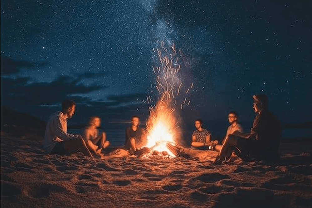 A group of people gathers around a campfire on the beach under a starry night sky.