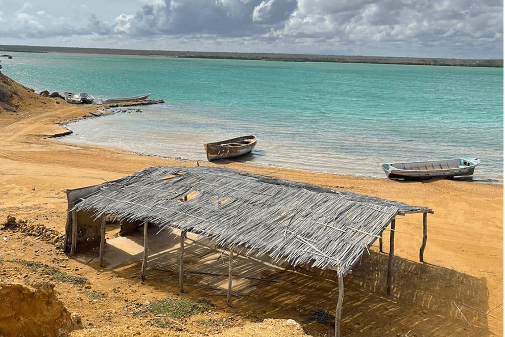 A thatched hut overlooks a sandy beach with several boats along a turquoise water shoreline.