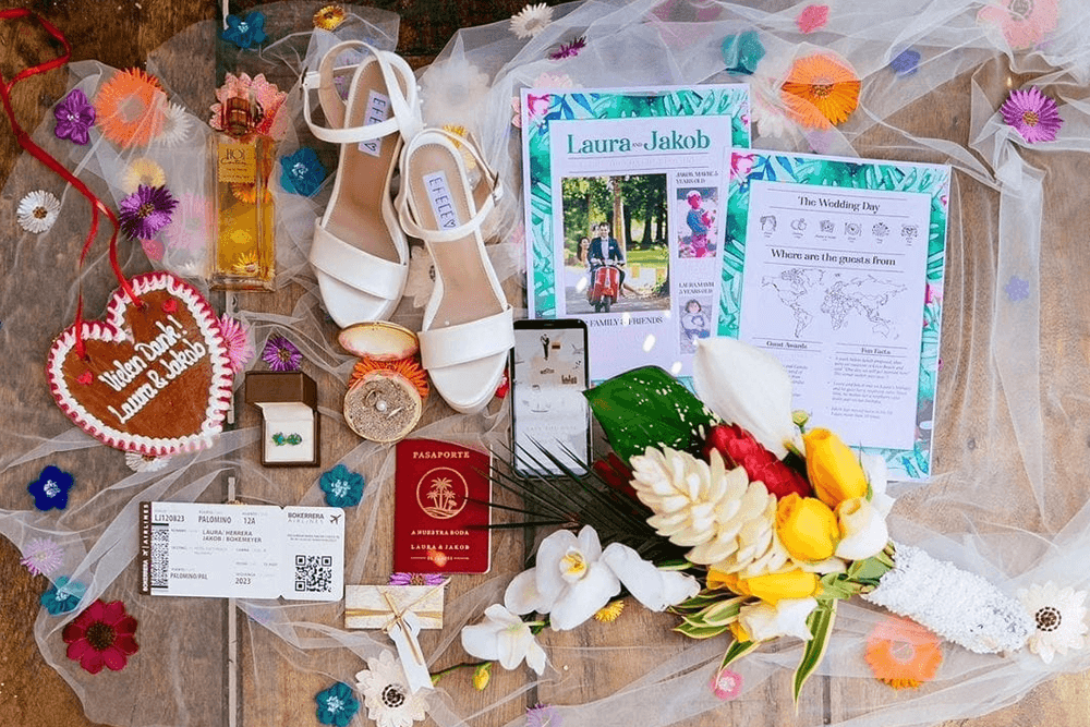 A wedding flat lay featuring shoes, a floral bouquet, a cake topper, invitations, a perfume bottle, and travel documents.