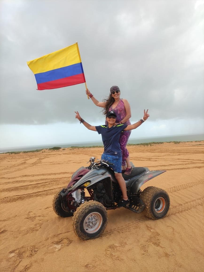 Two people pose on an ATV in a sandy landscape, with one holding a Colombian flag.
