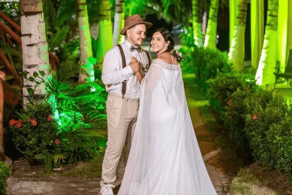A smiling couple in wedding attire stands together in a lush, green outdoor setting.