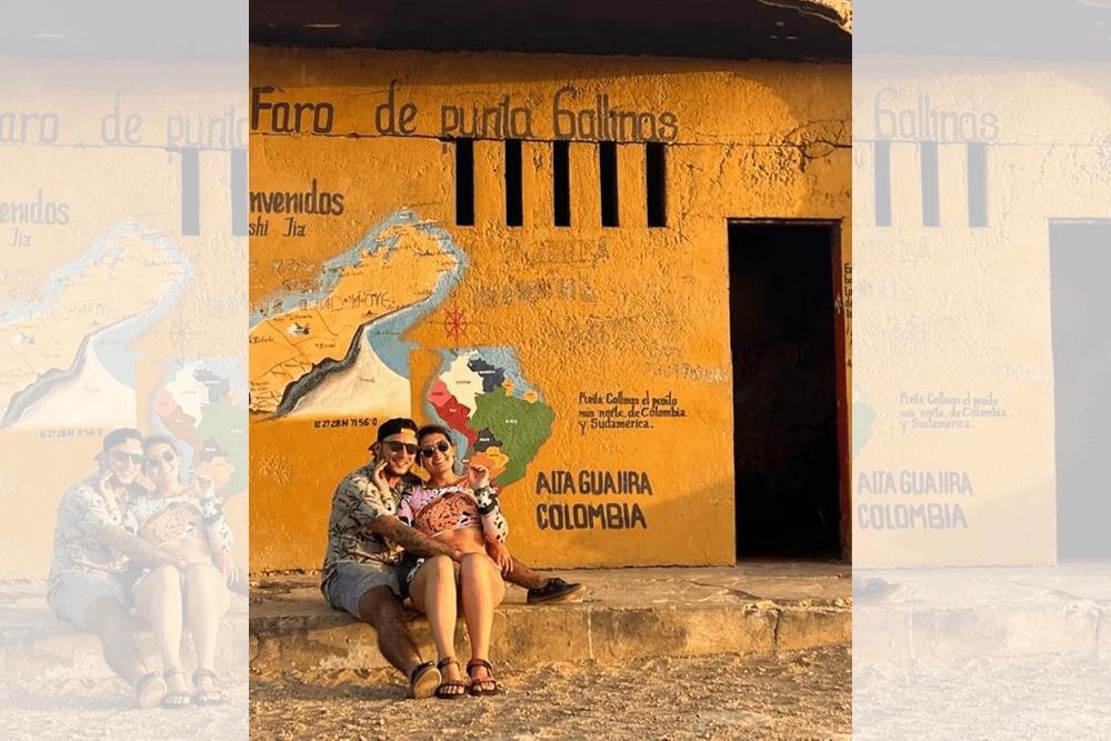 A couple sits together in front of a vibrant orange wall adorned with a map and text related to La Guajira, Colombia.