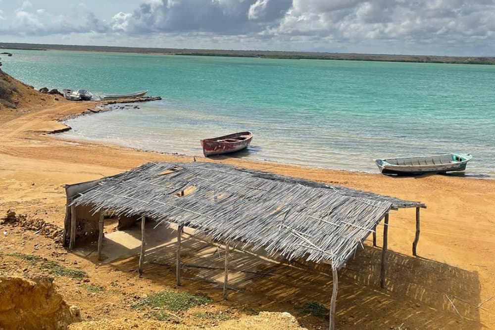 A thatched shelter on a sandy beach overlooking turquoise waters and several boats.
