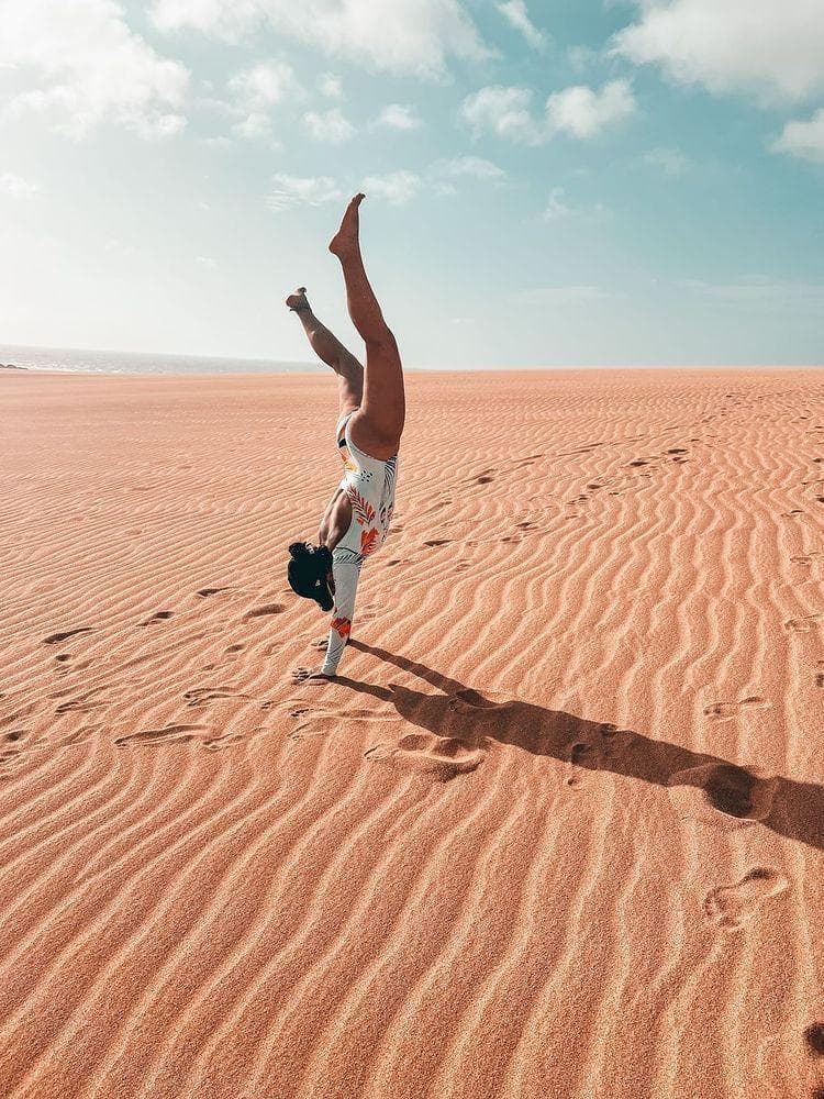 A person does a handstand on a wavy sand dune under a blue sky.