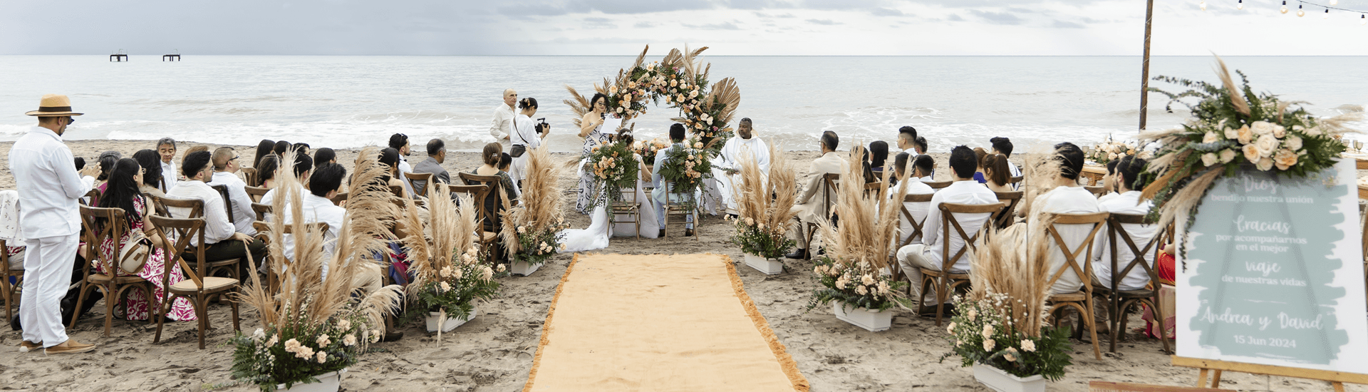 A beach wedding ceremony set against a backdrop of the ocean, featuring guests seated in rustic chairs and a floral arch.