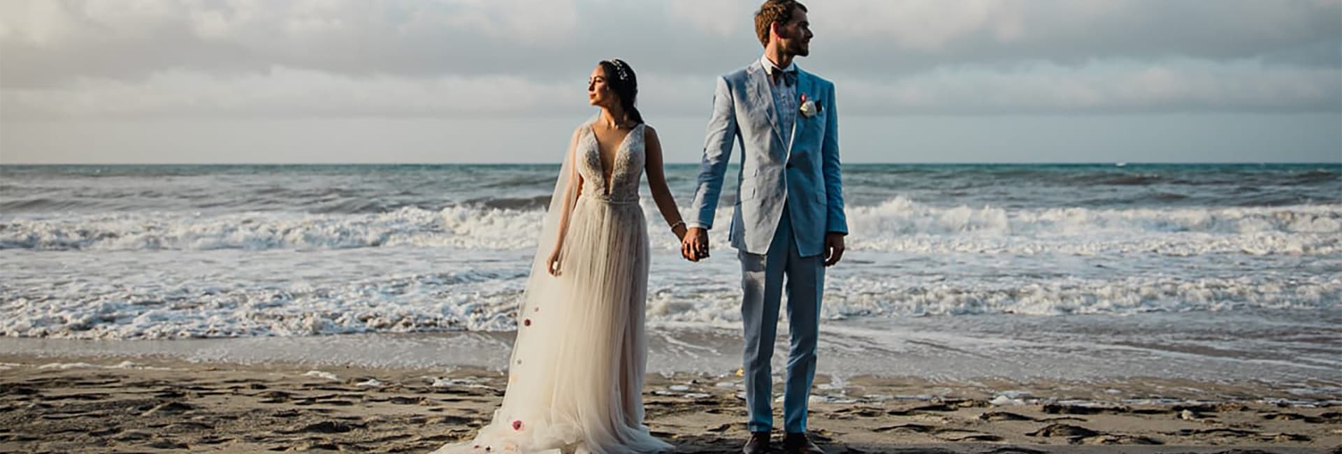 A bride and groom stand hand-in-hand on a beach, looking out at the ocean.