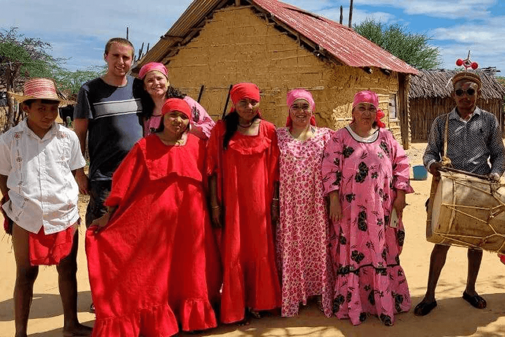 A group of people dressed in vibrant traditional clothing poses in front of a thatched-roof building.