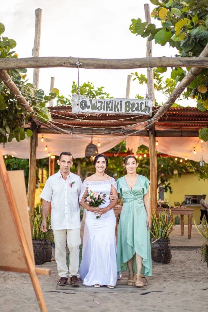 A bride in a white dress holds a bouquet, standing between her father and a woman in green, under a sign that reads "Waikiki Beach."