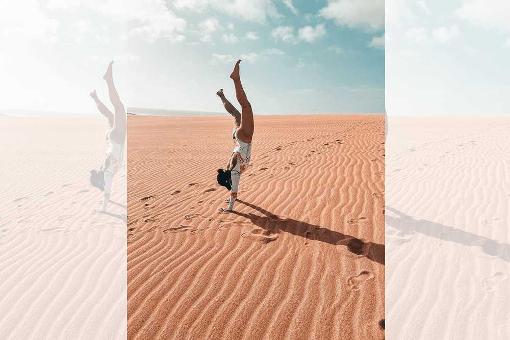 A person performs a handstand on a sandy beach under a bright sky.