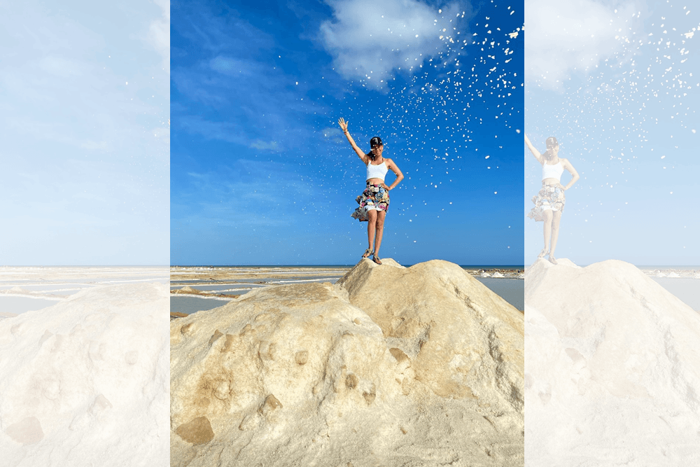 A person stands on a rock at the beach, playfully waving as confetti rains down against a bright blue sky.