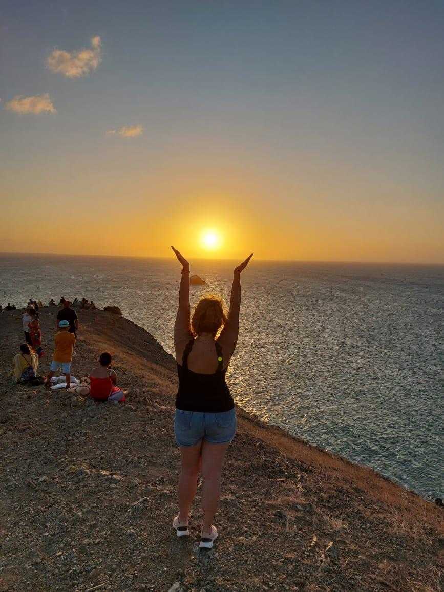 A person with raised arms stands on a rocky cliff at sunset overlooking the sea, surrounded by other spectators.