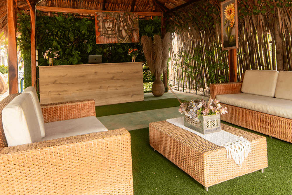 A cozy, open space featuring wicker furniture, a decorative coffee table, and a reception desk surrounded by lush greenery.