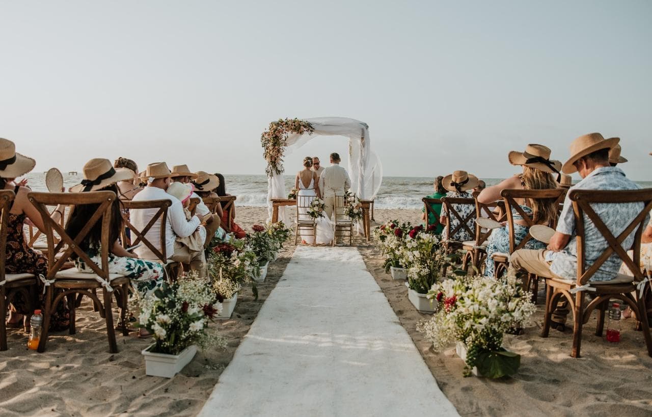 A couple stands under a floral arch at a beach wedding, surrounded by guests seated in straw hats.